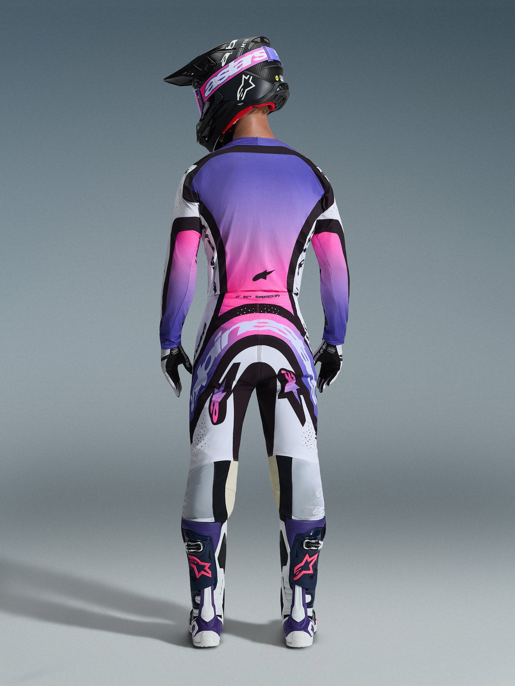 Widok z tyłu zawodnika motocrossowego w zestawie Alpinestars 2026 Supertech AFD Vista, koszulka i spodnie z gradientem bieli, fioletu i różu oraz czarnymi akcentami, czarny kask z fioletowymi goglami, fioletowo-białe buty, profesjonalny strój wyścigowy do jazdy w terenie.
