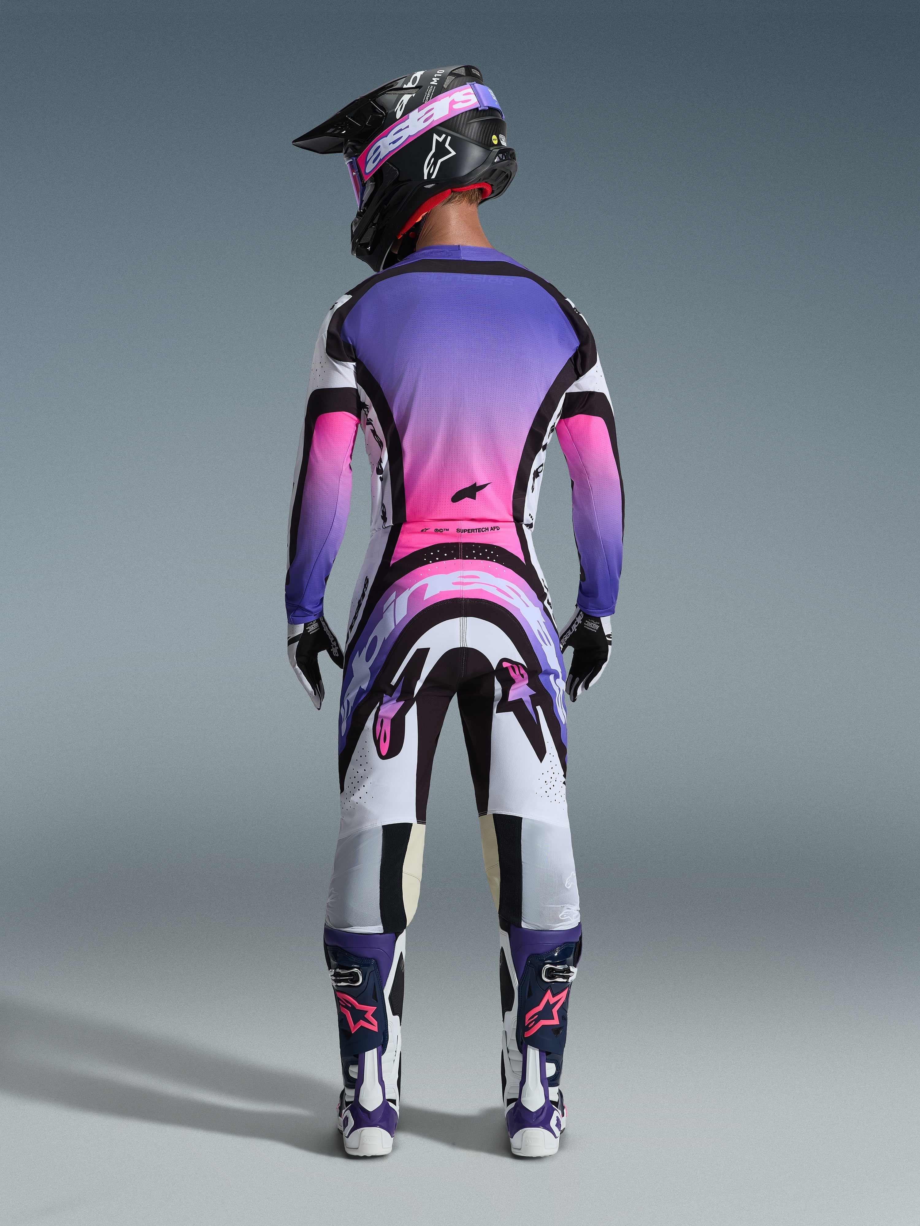 Widok z tyłu zawodnika motocrossowego w zestawie Alpinestars 2026 Supertech AFD Vista, koszulka i spodnie z gradientem bieli, fioletu i różu oraz czarnymi akcentami, czarny kask z fioletowymi goglami, fioletowo-białe buty, profesjonalny strój wyścigowy do jazdy w terenie.