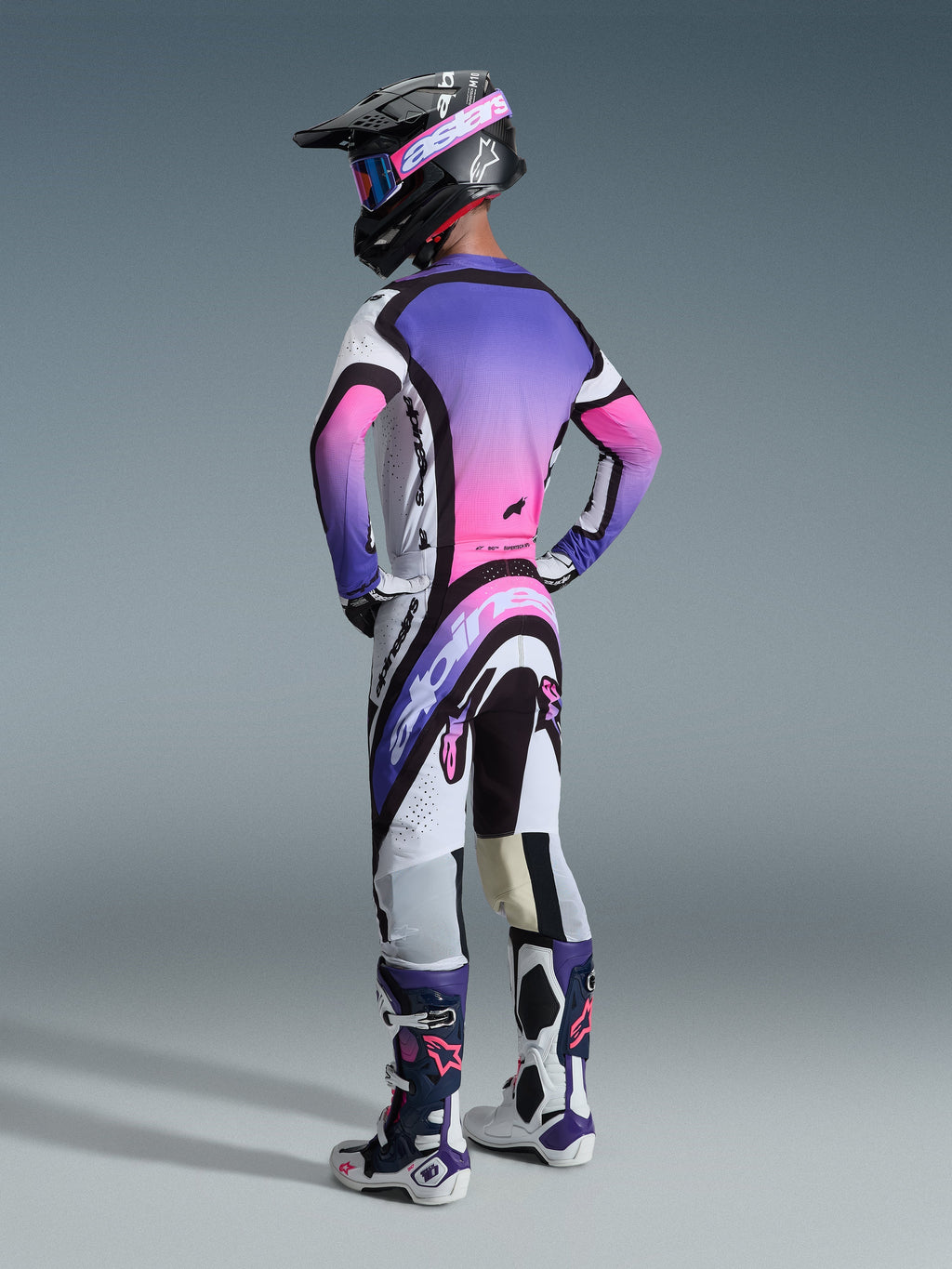 Motocrossista widoczny od tyłu ubrany w zestaw 2026 Supertech AFD Vista w kolorystyce White Purple Black, z koszulką o żywym fioletowo-różowym gradiencie na plecach, pasującymi spodniami z wyraźnym brandingiem Alpinestars, czarnym kaskiem z różowymi goglami oraz dopasowanymi biało-fioletowymi butami wyścigowymi.