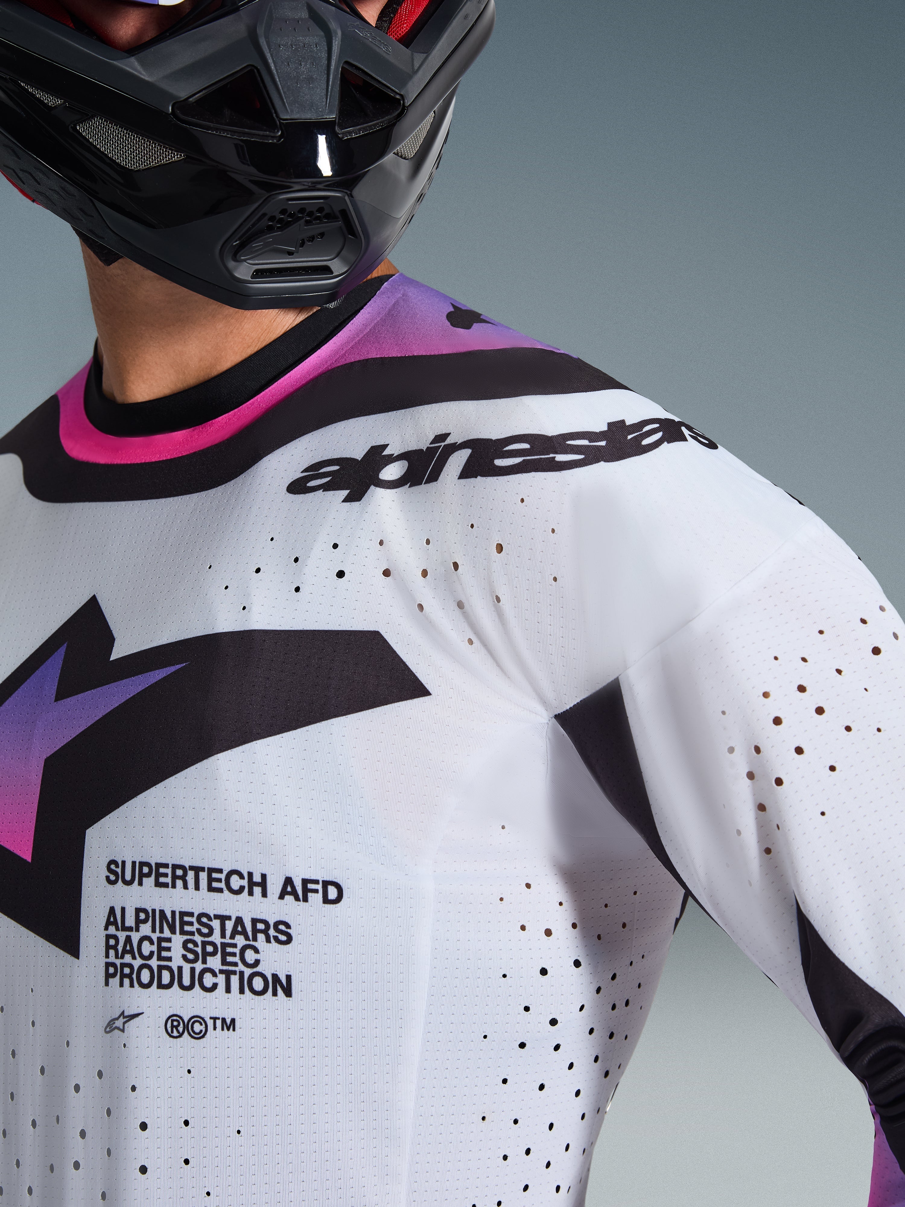 Zbliżenie na zawodnika motocrossowego w koszulce 2026 Supertech AFD Vista w kolorze biało-fioletowo-czarnym, z laserowo wycinanymi perforacjami dla wentylacji, napisem „SUPERTECH AFD” na klatce piersiowej i czarnym brandingiem Alpinestars na ramieniu, sportowa odzież wyścigowa off-road.