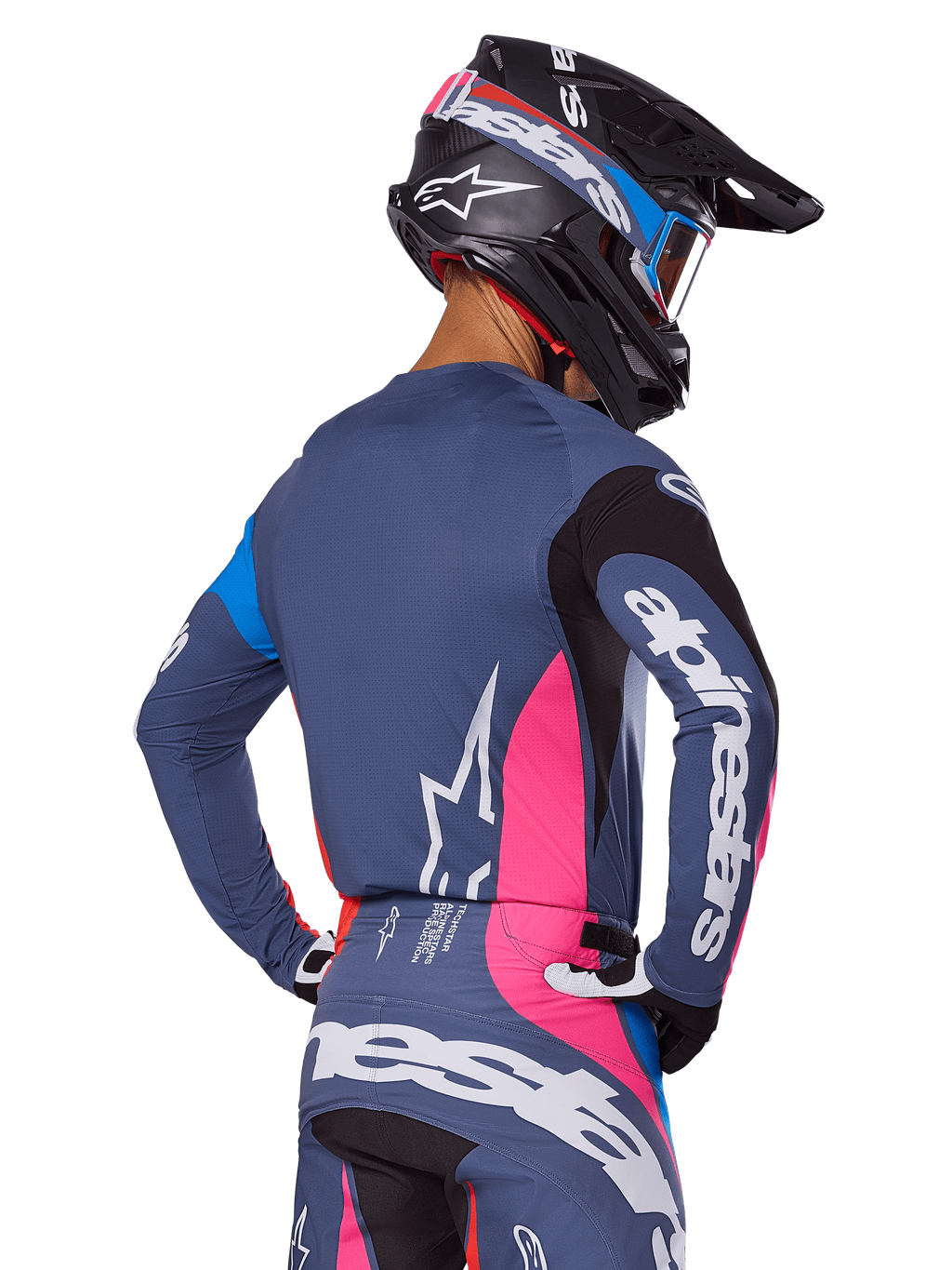 Motocyklista motocrossowy widziany od tyłu, ubrany w zestaw Alpinestars 2025 Techstar Dreem w kolorze szarym, niebiesko-szara koszulka i spodnie z różowymi i czarnymi panelami, białe logo Alpinestars na rękawach i plecach, czarny kask z niebiesko-różową grafiką.