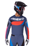 Alpinestars 2025 Techstar Dreem Jersey, koszulka motocrossowa, szara z niebieskimi, czerwonymi i różowymi akcentami, z laserowo wycinanymi perforacjami i dużym logo na klatce piersiowej, noszona z pasującymi spodniami i czarnym kaskiem wyścigowym