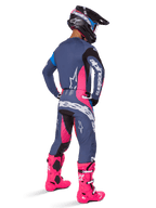 Zawodnik motocrossowy widoczny z tyłu, ubrany w zestaw odzieży Alpinestars 2025 Techstar Dreem, szary z jaskraworóżowymi i białymi akcentami, czarno-biały kask i jaskraworóżowe buty, kompletny strój wyścigowy do motocrossu.