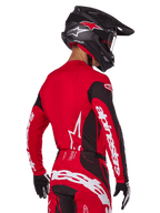 Zawodnik motocrossowy widziany z tyłu w zestawie Alpinestars 2025 Techstar Dreem, czerwono-czarna koszulka z białym brandingiem na rękawach, pasujące czerwono-czarne spodnie z wyraźnym białym logo na siedzeniu, czarny karbonowy kask z czerwonymi akcentami, czarno-białe rękawice, profesjonalny strój do wyścigów terenowych
