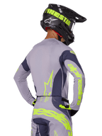 Zawodnik motocrossowy widoczny od tyłu w zestawie Alpinestars 2025 Techstar Dreem, szara koszulka i spodnie z jaskrawym neonowym żółto-zielonym brandingiem, pasujący czarny kask z neonową grafiką, profesjonalny strój wyścigowy