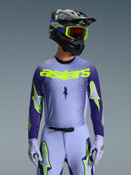 Alpinestars 2026 Supertech Scenz Jersey, koszulka motocrossowa, fioletowa i neonowo-żółta, noszona przez motocyklistę z dopasowanym czarno-fioletowym kaskiem, neonowo-żółtymi goglami i fioletowymi spodniami wyścigowymi, z neonowo-żółtym brandingiem astars na piersi i logo gwiazdy na rękawach.