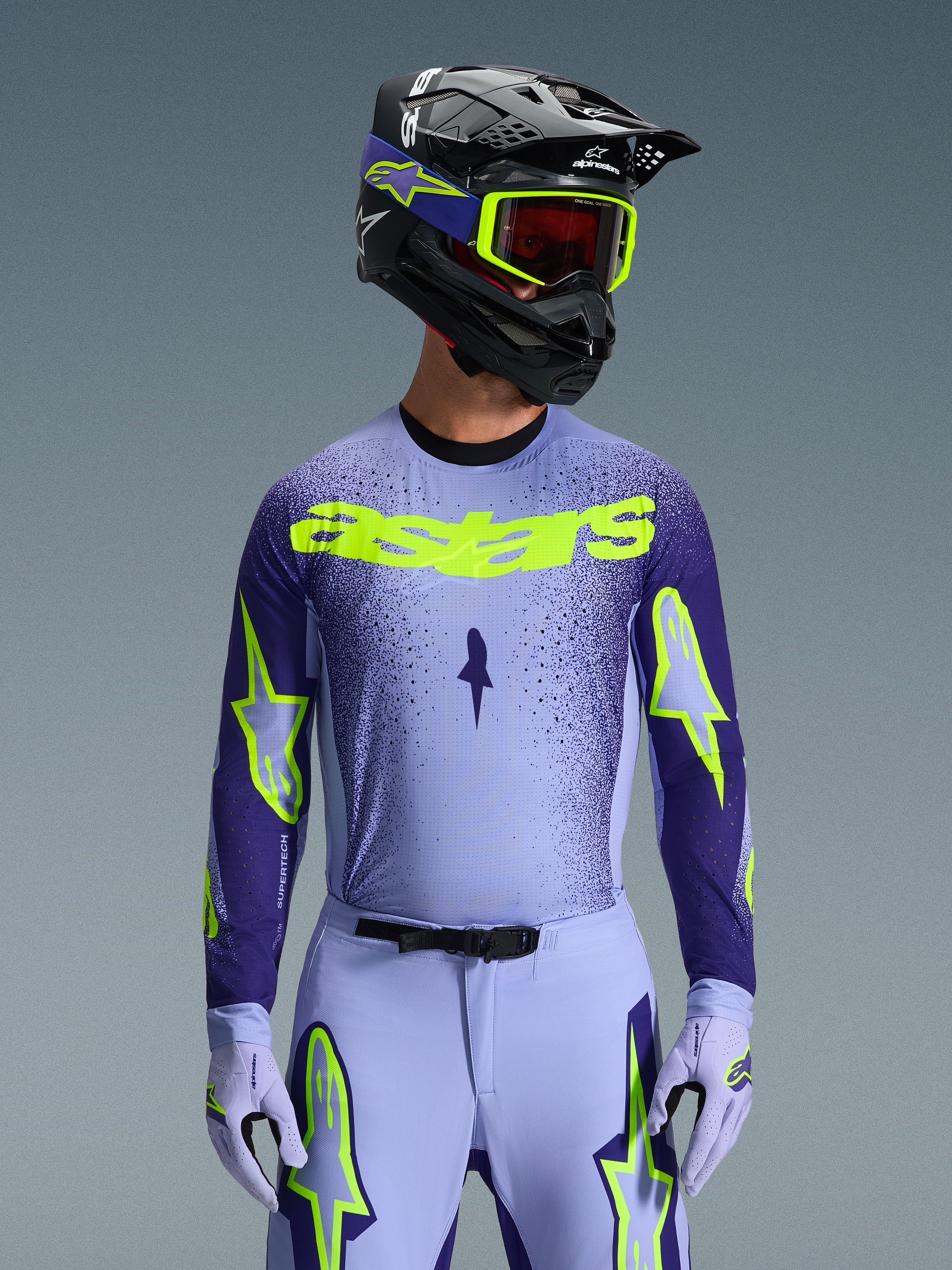Alpinestars 2026 Supertech Scenz Jersey, koszulka motocrossowa, fioletowa i neonowo-żółta, noszona przez motocyklistę z dopasowanym czarno-fioletowym kaskiem, neonowo-żółtymi goglami i fioletowymi spodniami wyścigowymi, z neonowo-żółtym brandingiem astars na piersi i logo gwiazdy na rękawach.