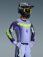 Kierowca motocrossowy w koszulce Alpinestars Supertech Scenz 2026, fioletowo-żółty fluo, dopasowane fioletowe spodnie z jaskrawymi akcentami, czarny kask z żółtymi goglami i fioletowe rękawice wyścigowe, profesjonalny zestaw odzieży do jazdy terenowej