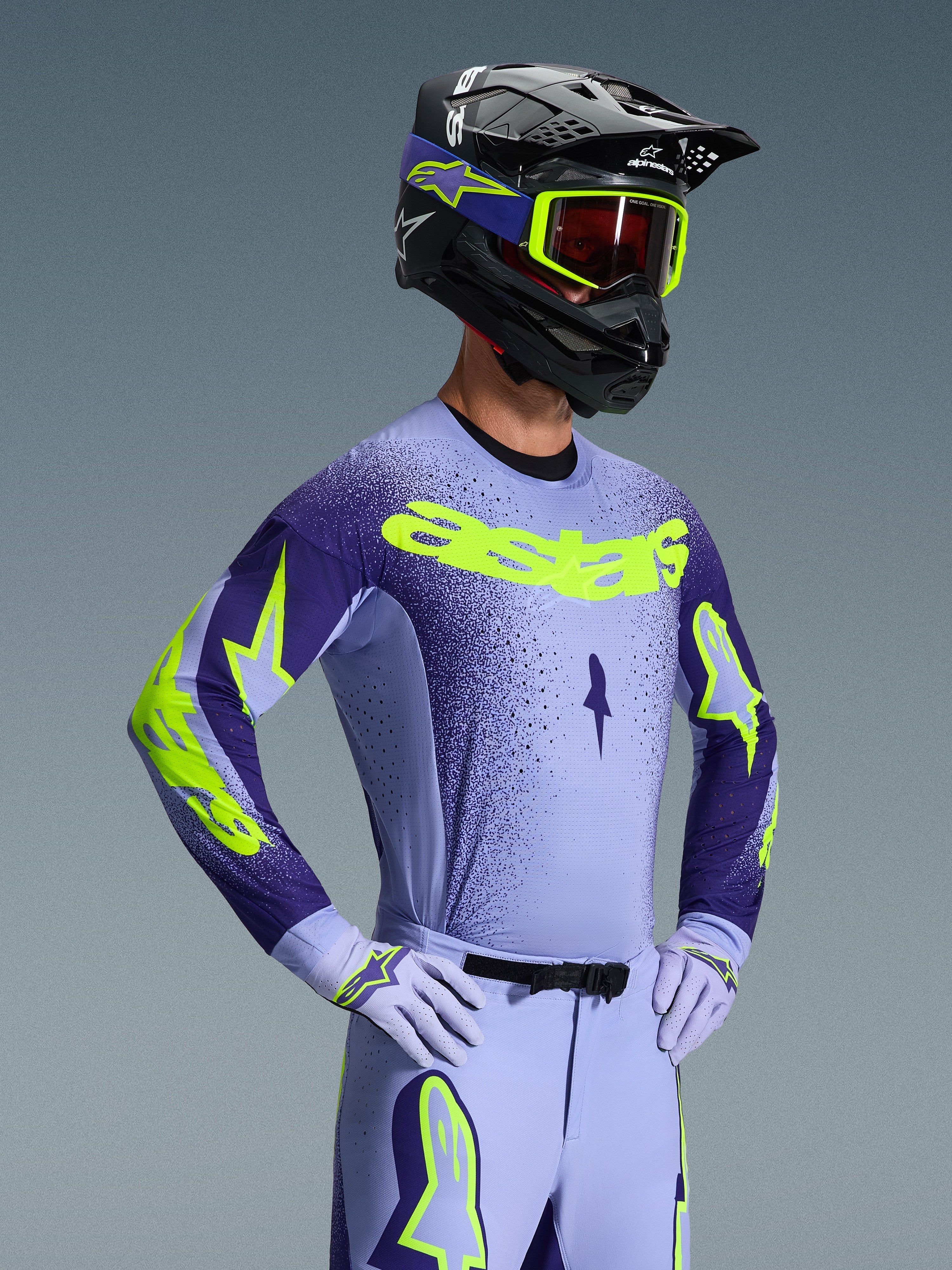 Kierowca motocrossowy w koszulce Alpinestars Supertech Scenz 2026, fioletowo-żółty fluo, dopasowane fioletowe spodnie z jaskrawymi akcentami, czarny kask z żółtymi goglami i fioletowe rękawice wyścigowe, profesjonalny zestaw odzieży do jazdy terenowej