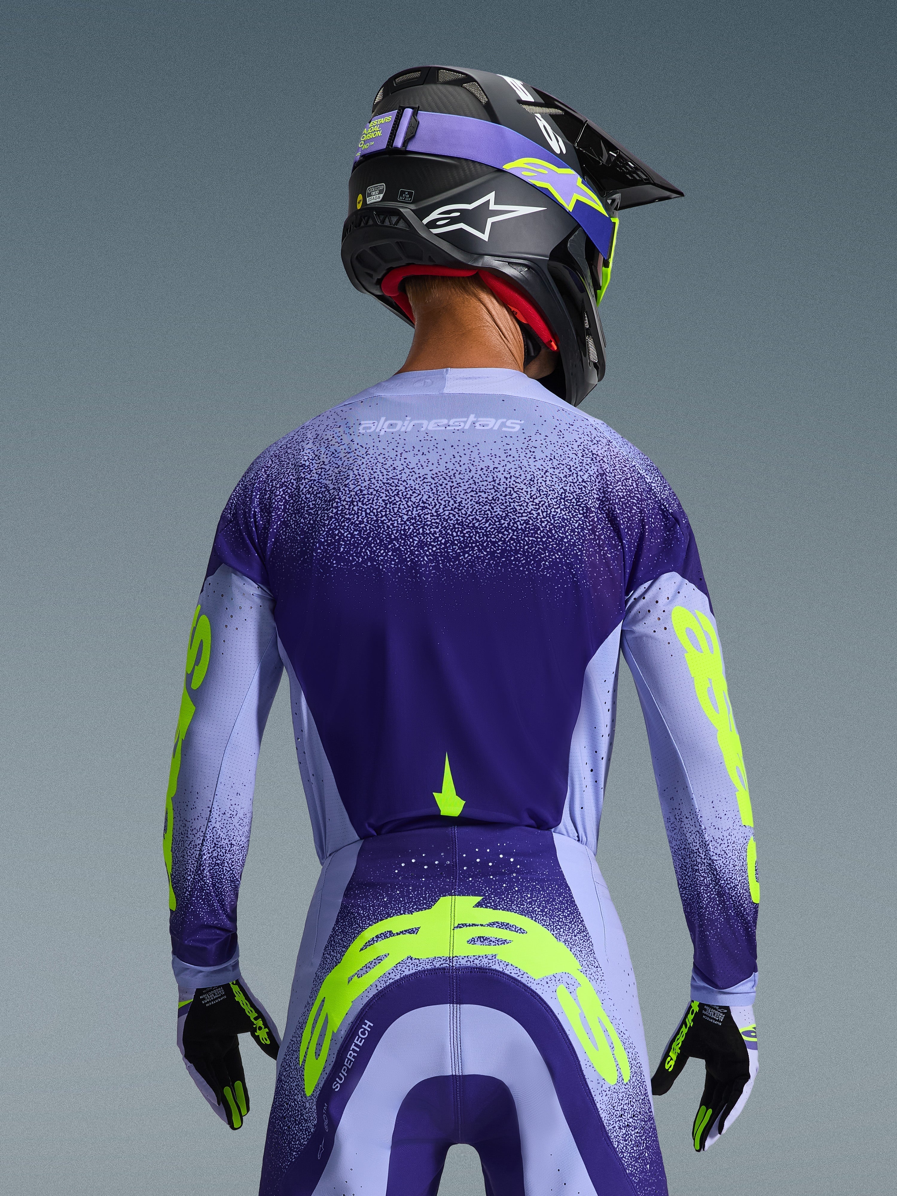Koszulka Alpinestars Supertech Scenz 2026, fioletowa i jasnoniebieska z jaskrawożółtymi akcentami, widok z tyłu na zawodniku w dopasowanych spodniach motocrossowych, czarnym kasku i rękawicach, profesjonalna odzież do wyścigów terenowych
