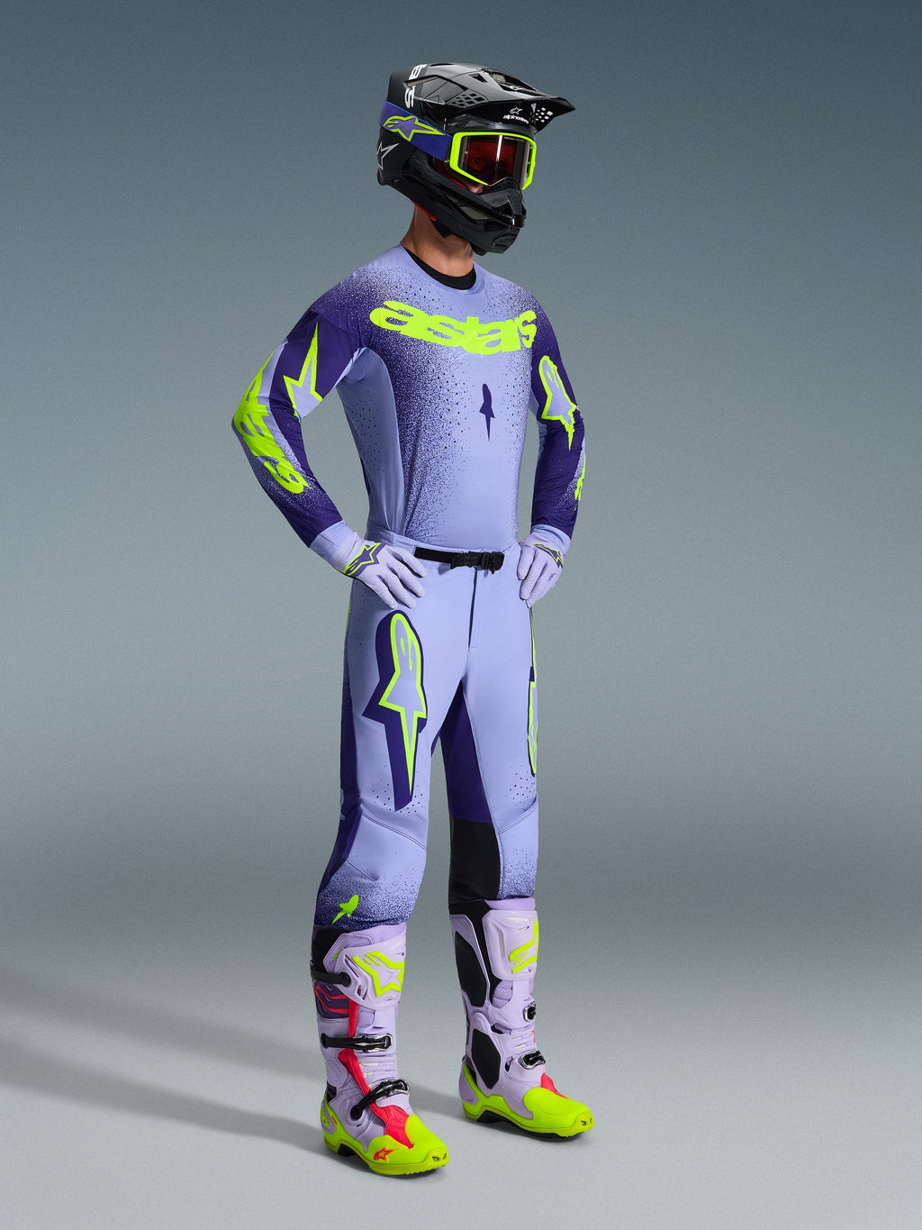 Zawodnik motocrossowy w koszulce 2026 Supertech Scenz i pełnym zestawie wyścigowym, fioletowo-żółty design Fluo z jasnofioletową bazą i neonowymi akcentami, pasujące spodnie z wyrazistym logo gwiazdy, czarny kask z neonowo-żółtymi goglami, dopasowane fioletowe i neonowo-żółte buty, profesjonalny zestaw odzieży off-road