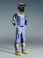 Zawodnik motocrossowy w koszulce 2026 Supertech Scenz i pełnym zestawie wyścigowym, fioletowo-żółty design Fluo z jasnofioletową bazą i neonowymi akcentami, pasujące spodnie z wyrazistym logo gwiazdy, czarny kask z neonowo-żółtymi goglami, dopasowane fioletowe i neonowo-żółte buty, profesjonalny zestaw odzieży off-road
