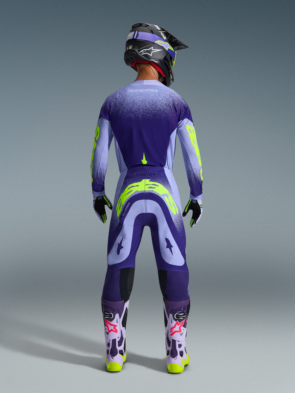 Motocrossowiec widoczny od tyłu w koszulce Alpinestars 2026 Supertech Scenz w kolorze Purple Yellow Fluo, z fioletowym gradientem i żółtymi detalami marki, w komplecie z dopasowanymi spodniami motocrossowymi i fioletowymi butami, pełny zestaw wyścigowy off-road