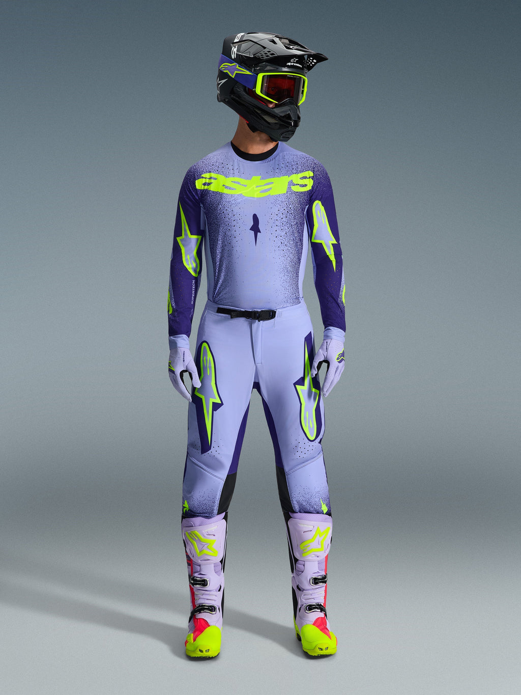 Motocyklista motocrossowy ubrany w pełny strój wyścigowy Alpinestars 2026 Supertech Scenz, fioletowo-neonowożółta koszulka i pasujące spodnie, czarny kask z żółtymi goglami, jasnofioletowe i neonowe buty, profesjonalny strój o sportowym kroju do wyścigów off-road
