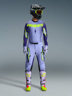 Motocyklista motocrossowy ubrany w pełny strój wyścigowy Alpinestars 2026 Supertech Scenz, fioletowo-neonowożółta koszulka i pasujące spodnie, czarny kask z żółtymi goglami, jasnofioletowe i neonowe buty, profesjonalny strój o sportowym kroju do wyścigów off-road