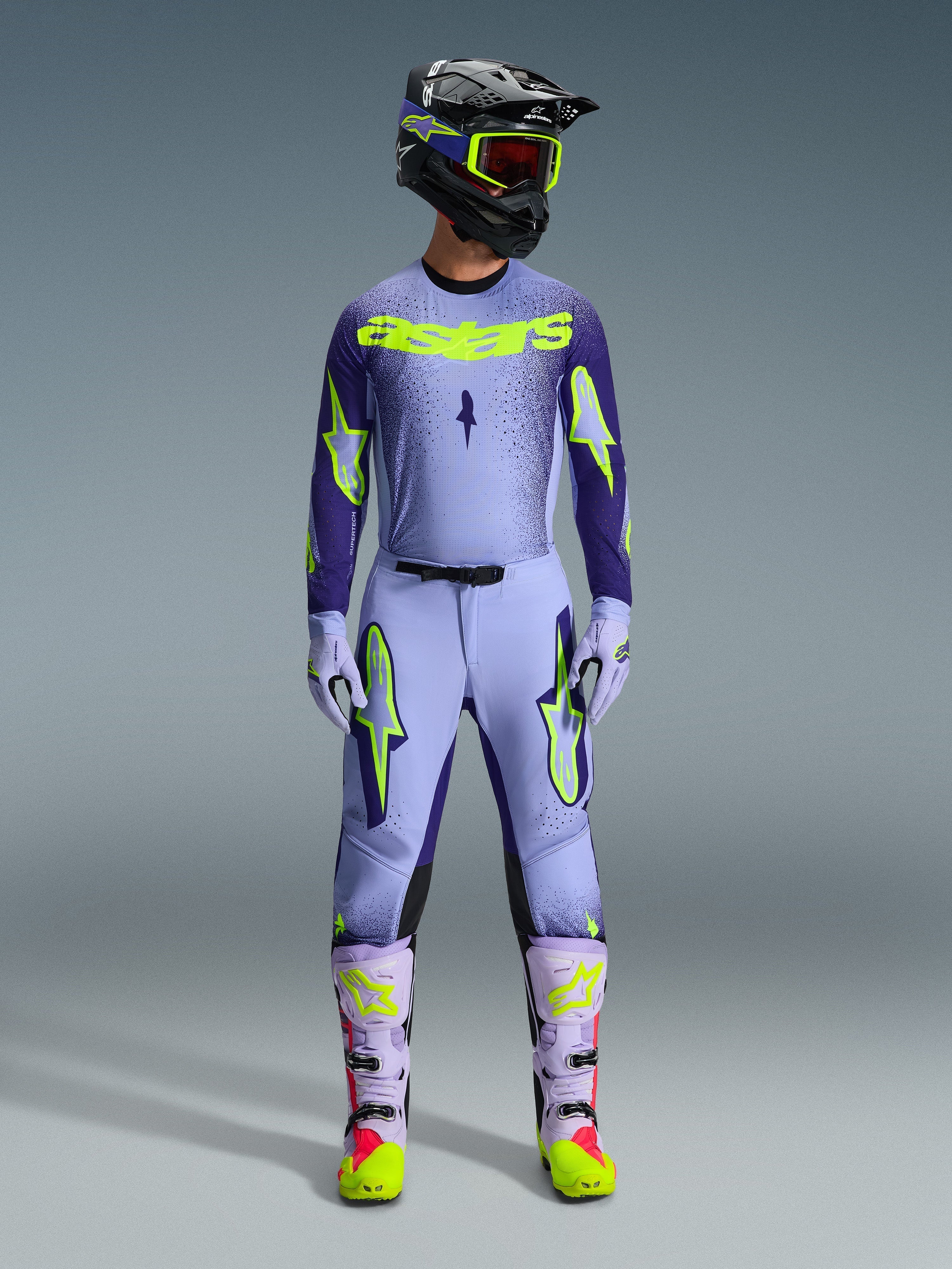 Motocyklista motocrossowy ubrany w pełny strój wyścigowy Alpinestars 2026 Supertech Scenz, fioletowo-neonowożółta koszulka i pasujące spodnie, czarny kask z żółtymi goglami, jasnofioletowe i neonowe buty, profesjonalny strój o sportowym kroju do wyścigów off-road