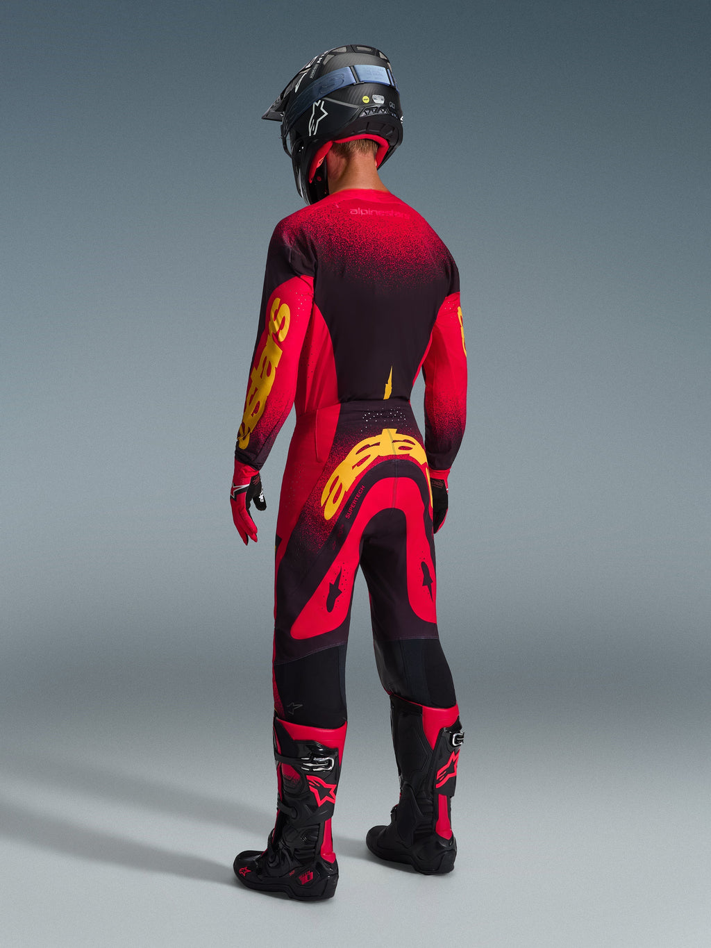 Zawodnik motocrossowy Alpinestars widziany od tyłu, pełny strój wyścigowy, czerwona koszulka z czarnymi i żółtymi akcentami, pasujące czerwono-czarne spodnie z dużym żółtym logo Alpinestars, czarny kask, czarno-czerwone buty i rękawice, profesjonalny zestaw off-road