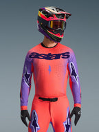 Alpinestars Supertech Scenz Jersey 2026, koszulka motocrossowa, koralowa i fioletowa, z dużym logo „astars” i wzorami gwiazd na rękawach, noszona z dopasowanymi spodniami i wielokolorowym kaskiem, zaawansowana odzież terenowa