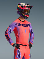 Zawodnik motocrossowy w koszulce Alpinestars 2026 Supertech Scenz, kolory Hot Coral i fioletowy z wyraźnym brandingiem „astars” na klatce piersiowej, dopasowany zestaw z koralowymi spodniami i fioletowo-koralowym kaskiem, laserowo wycinane perforacje dla wysokiej wydajności w wyścigach terenowych.