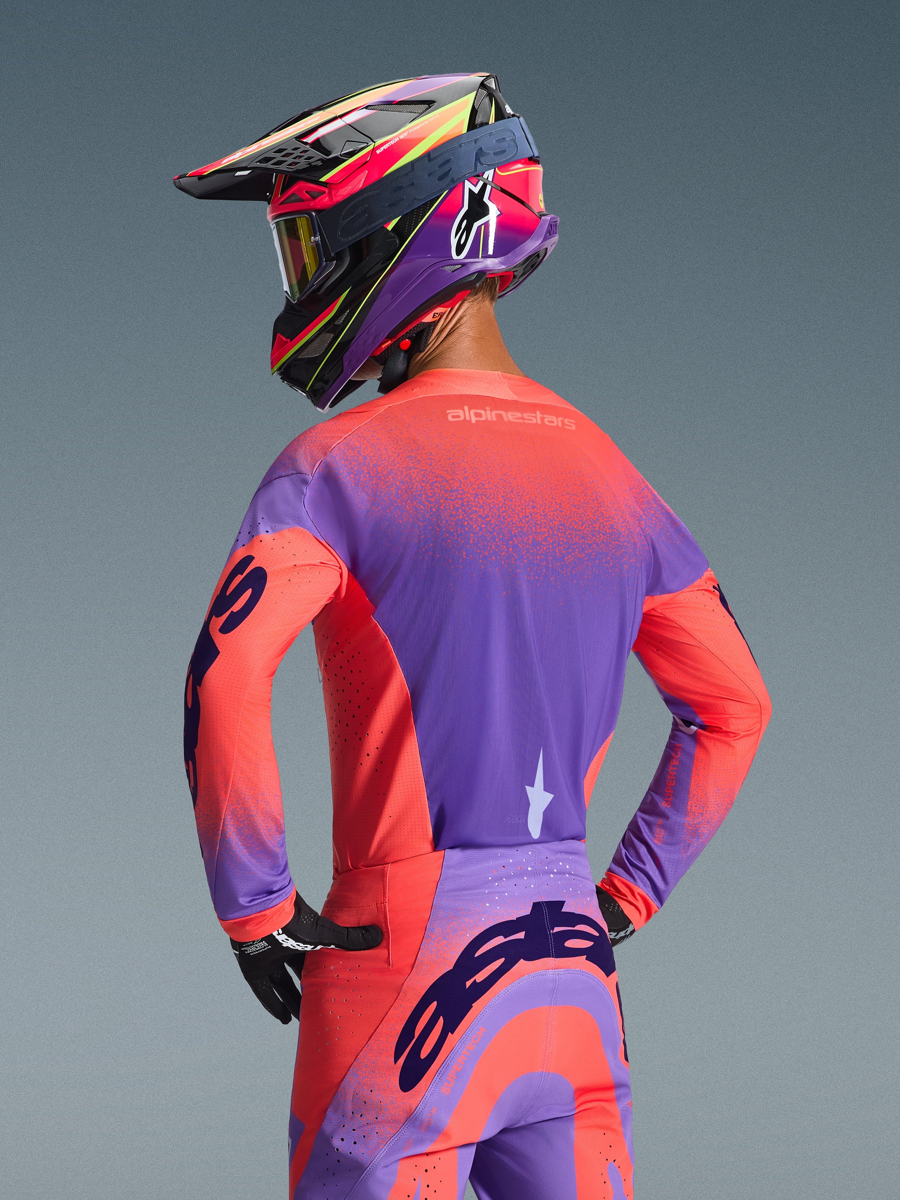 Motocyklista motocrossowy widoczny od tyłu w koszulce Alpinestars Supertech Scenz 2026 w kolorach Hot Coral i Purple, z żywym nakrapianym wzorem gradientowym, w zestawie z dopasowanymi spodniami motocrossowymi i kolorowym kaskiem wyścigowym, profesjonalny sprzęt off-road