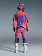 Motocyklista motocrossowy widziany od tyłu w zestawie Supertech Scenz 2026 w kolorze Hot Coral Purple, wyrazista koszulka i spodnie z gradientem, logo Alpinestars, dopasowany kask, biało-czarne buty i czarne rękawice, profesjonalny sprzęt do wyścigów terenowych.