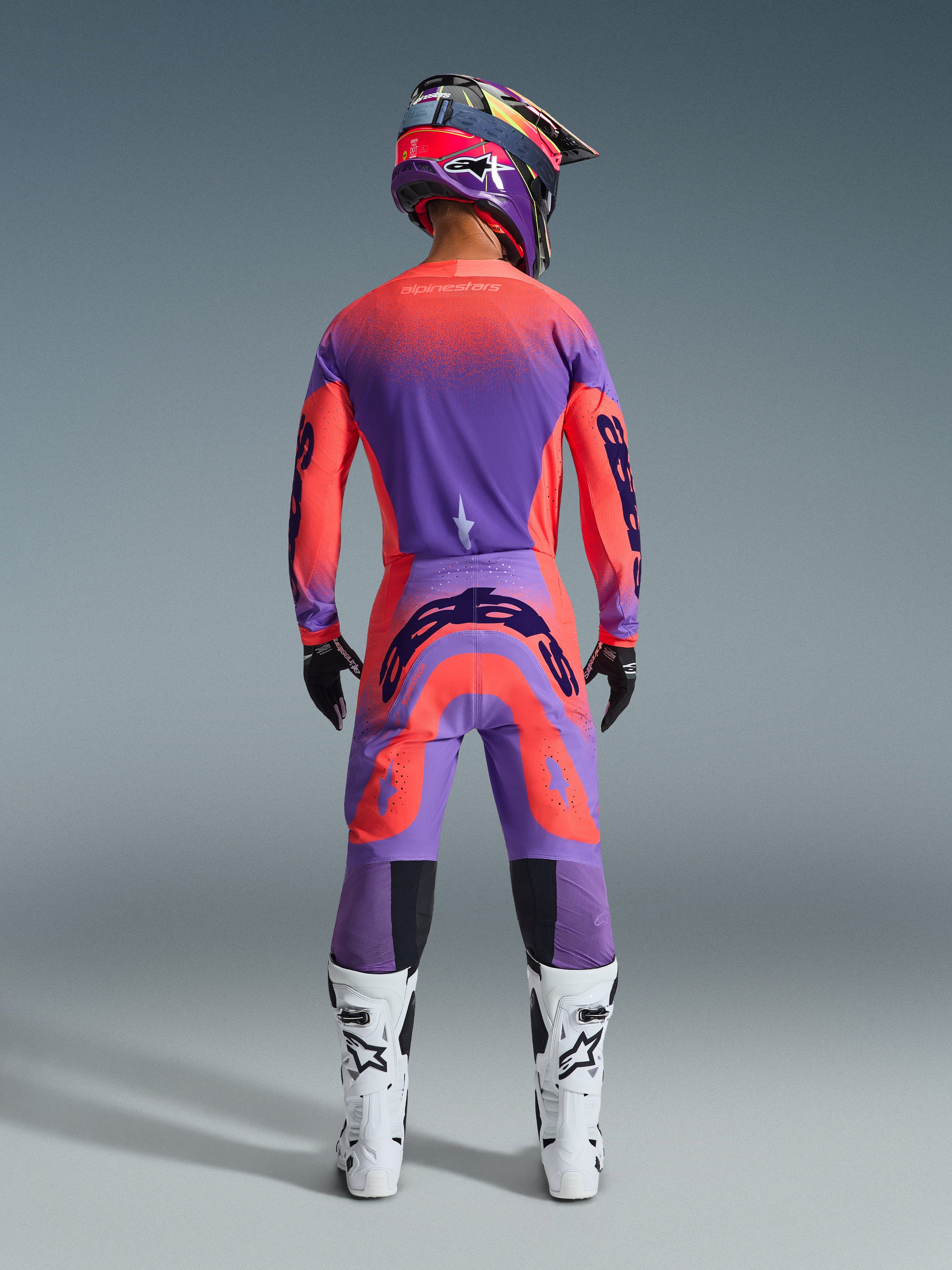 Motocyklista motocrossowy widziany od tyłu w zestawie Supertech Scenz 2026 w kolorze Hot Coral Purple, wyrazista koszulka i spodnie z gradientem, logo Alpinestars, dopasowany kask, biało-czarne buty i czarne rękawice, profesjonalny sprzęt do wyścigów terenowych.