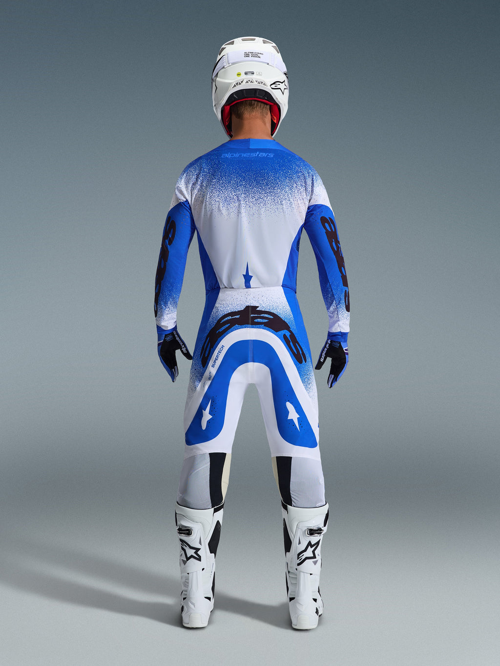 Zawodnik motocrossowy widziany z tyłu w zestawie Alpinestars 2026 Supertech Scenz w kolorze Ucla Blue White, niebiesko-biała koszulka z czarnym brandingiem, pasujące spodnie, biały kask i białe buty, profesjonalny sprzęt wyścigowy do motocrossu off-road