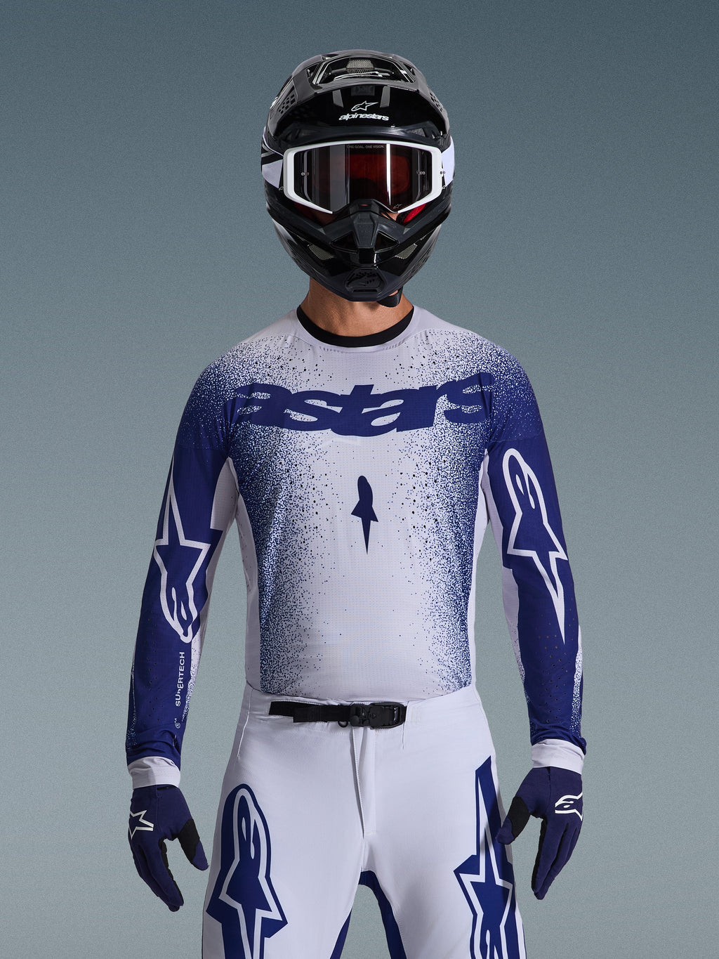 Zawodnik motocrossowy w koszulce Alpinestars 2026 Supertech Scenz, jasnoszara i ciemnoniebieska z nakrapianym wzorem gradientowym i wyraźnym brandingiem „astars”. Noszona w zestawie z dopasowanymi białymi spodniami motocrossowymi, ciemnoniebieskimi rękawicami i czarnym kaskiem z goglami; profesjonalny strój do wyścigów terenowych.