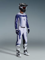 Motocyklista motocrossowy w kompletnym profesjonalnym stroju wyścigowym, koszulka 2026 Supertech Scenz w kolorze jasnoszarym i ciemnoniebieskim, pasujące spodnie motocrossowe z grafiką gwiazdy, czarny kask z czerwonymi goglami, biało-czarne buty wyczynowe, dopasowane niebiesko-białe rękawice do wyścigów terenowych