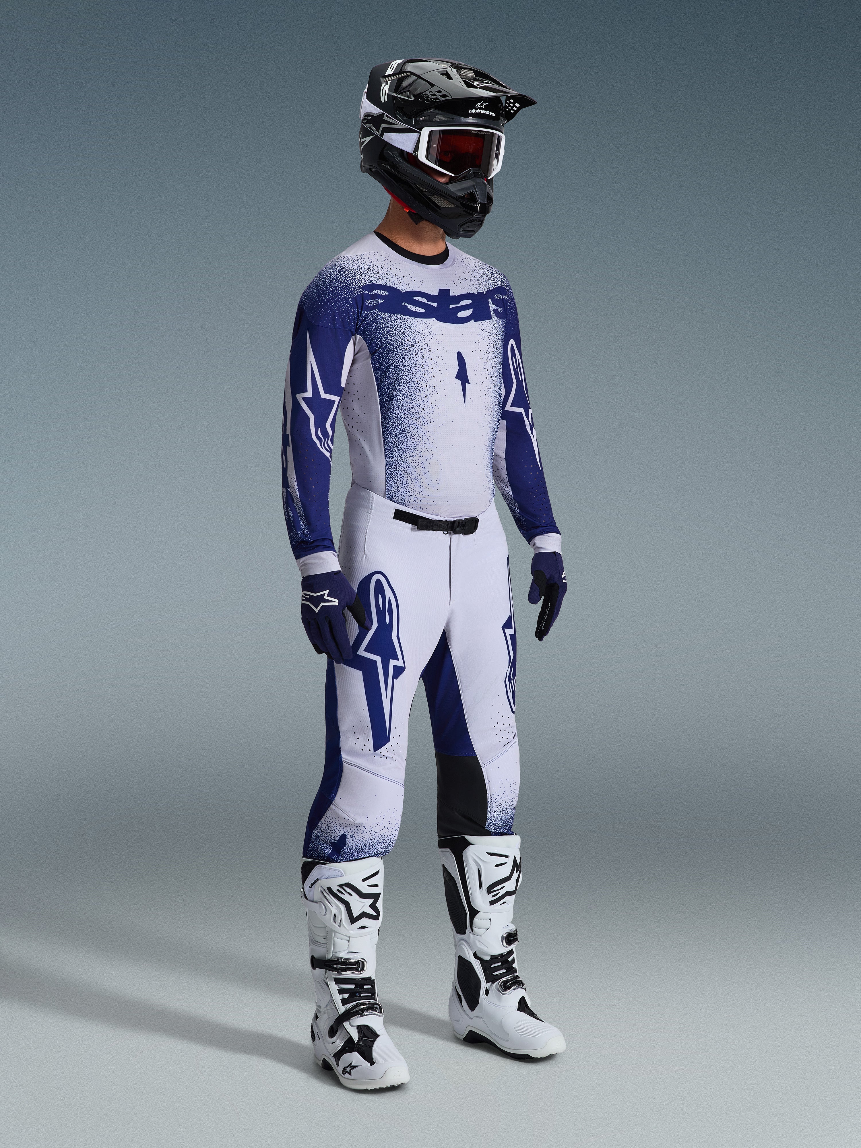 Motocyklista motocrossowy w kompletnym profesjonalnym stroju wyścigowym, koszulka 2026 Supertech Scenz w kolorze jasnoszarym i ciemnoniebieskim, pasujące spodnie motocrossowe z grafiką gwiazdy, czarny kask z czerwonymi goglami, biało-czarne buty wyczynowe, dopasowane niebiesko-białe rękawice do wyścigów terenowych