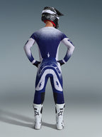 Motocrossista widziany od tyłu w koszulce Alpinestars 2026 Supertech Scenz, jasnoszaro-ciemnoniebieskiej z kropkowanym wzorem gradientowym, pasujące spodnie motocrossowe, białe buty wyścigowe i czarny kask, kompletny strój wyścigowy off-road