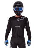 Alpinestars 2025 Racer Graphite Jersey, odzież motocrossowa, czarna z szarymi logotypami, noszona z pasującym czarnym kaskiem, niebieskimi goglami i niebiesko-szarymi rękawicami, dopasowany krój sportowy z logo na piersi i rękawach