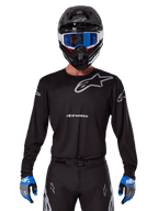 Alpinestars 2025 Racer Graphite Jersey, odzież motocrossowa, czarna z szarymi logotypami, noszona z pasującym czarnym kaskiem, niebieskimi goglami i niebiesko-szarymi rękawicami, dopasowany krój sportowy z logo na piersi i rękawach
