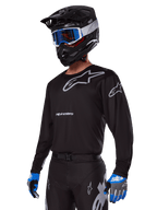 Zawodnik motocrossowy w zestawie Alpinestars 2025 Racer Graphite, czarna koszulka z szarymi logotypami Alpinestars, pasujące czarne spodnie z szarymi akcentami, czarny kask z niebiesko-białymi goglami, niebiesko-szare rękawice, nowoczesny strój techniczny do jazdy off-road