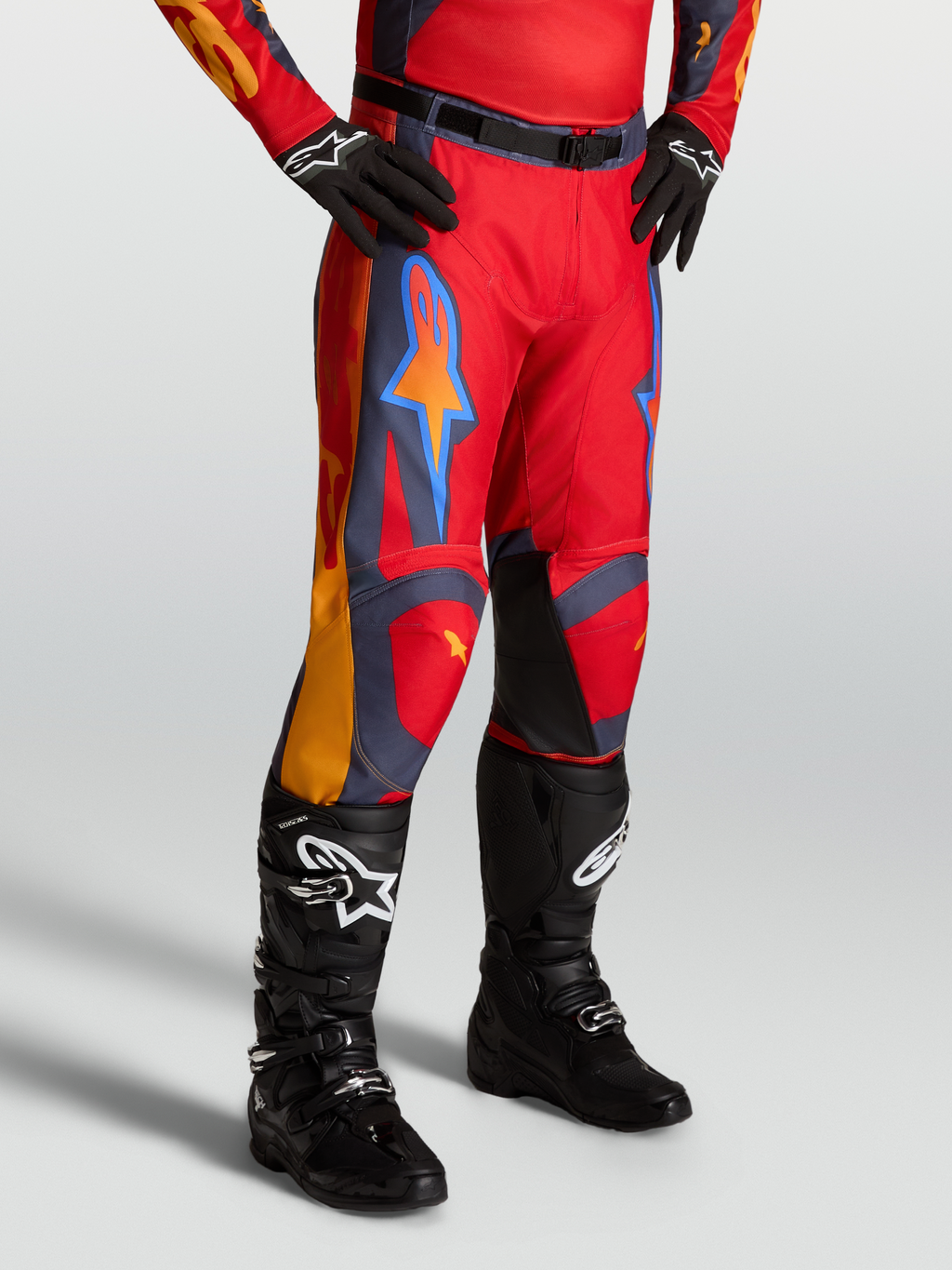 Alpinestars Spodnie Racer Hunt, czerwone spodnie motocrossowe z szarymi i koralowymi akcentami, na modelu z czarnymi butami.