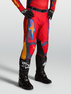 Alpinestars Spodnie Racer Hunt, czerwone spodnie motocrossowe z szarymi i koralowymi akcentami, na modelu z czarnymi butami.