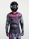 Alpinestars Koszulka Racer Lone, męska koszulka motocrossowa, szary wzór camo i fioletowe detale, lekka techniczna tkanina, prezentowana na modelu w kasku i goglach.