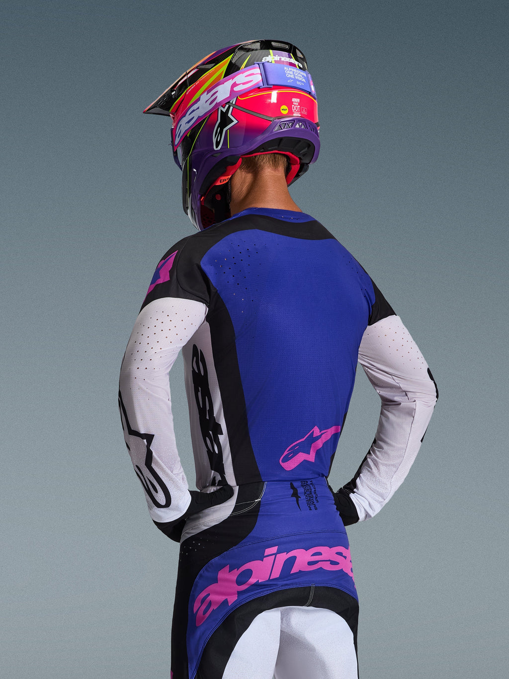 Zawodnik motocrossowy od tyłu w koszulce 2026 Techstar Knif, czarny, jasnoszary i fioletowy, białe perforowane rękawy i różowe logo Alpinestars, dopasowany fioletowo-różowy kask, profesjonalny strój do wyścigów off-road