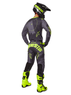 Widok z tyłu zawodnika motocrossowego w zestawie Racer Hollow 2025 w kolorze Dark Camo Yellow Fluo, szara koszulka moro z jaskrawożółtymi akcentami, pasujące spodnie z wyraźnym logo na siedzeniu, jaskrawożółte buty fluo i czarny kask z żółtymi goglami.