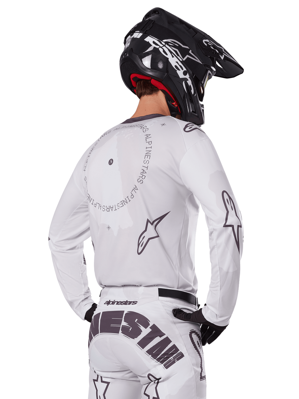 Zawodnik motocrossowy widoczny od tyłu w białym zestawie Alpinestars 2025 Racer Hollow, biała koszulka z długim rękawem z szarymi logo gwiazdy i okrągłymi grafikami, pasujące białe spodnie z wyraźnym brandingiem na siedzeniu, czarny kask, profesjonalny strój off-road