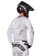 Zawodnik motocrossowy widoczny od tyłu w białym zestawie Alpinestars 2025 Racer Hollow, biała koszulka z długim rękawem z szarymi logo gwiazdy i okrągłymi grafikami, pasujące białe spodnie z wyraźnym brandingiem na siedzeniu, czarny kask, profesjonalny strój off-road