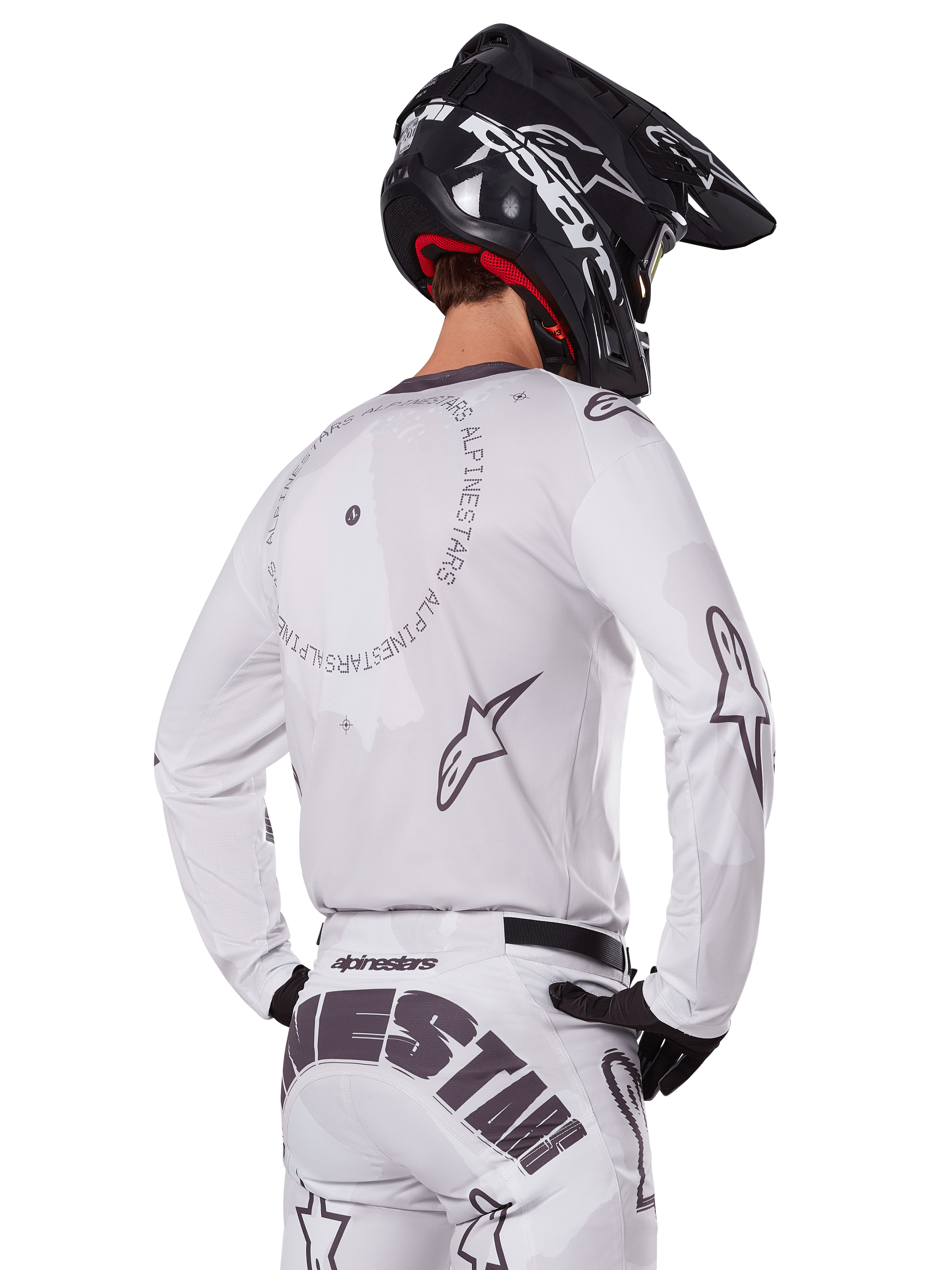 Zawodnik motocrossowy widoczny od tyłu w białym zestawie Alpinestars 2025 Racer Hollow, biała koszulka z długim rękawem z szarymi logo gwiazdy i okrągłymi grafikami, pasujące białe spodnie z wyraźnym brandingiem na siedzeniu, czarny kask, profesjonalny strój off-road