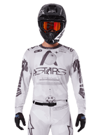 Alpinestars 2025 Racer Hollow Jersey, biało-czarna koszulka motocrossowa, noszona z pasującymi białymi spodniami MX i czarnym kaskiem z pomarańczowymi goglami, z wyraźnym brandingiem Alpinestars i grafikami z motywem globu, profesjonalna odzież wyścigowa off-road