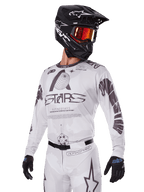 Zestaw Alpinestars 2025 Racer Hollow, koszulka i spodnie motocrossowe, białe z czarnymi i szarymi grafikami, z brandingiem „ASTARS” i motywami ażurowego globu, w połączeniu z czarnym kaskiem, czerwonymi lustrzanymi goglami i czarnymi rękawicami, zaprojektowany do wyczynowej jazdy off-road