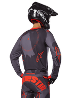 Zawodnik motocrossowy Alpinestars od tyłu w pełnym stroju wyścigowym, koszulka Racer Hollow w szarym kamuflażu z jaskrawopomarańczowymi akcentami i okrągłym logo na plecach, pasujące spodnie z dużym pomarańczowym napisem Alpinestars, czarny kask i pomarańczowo-czarne rękawice