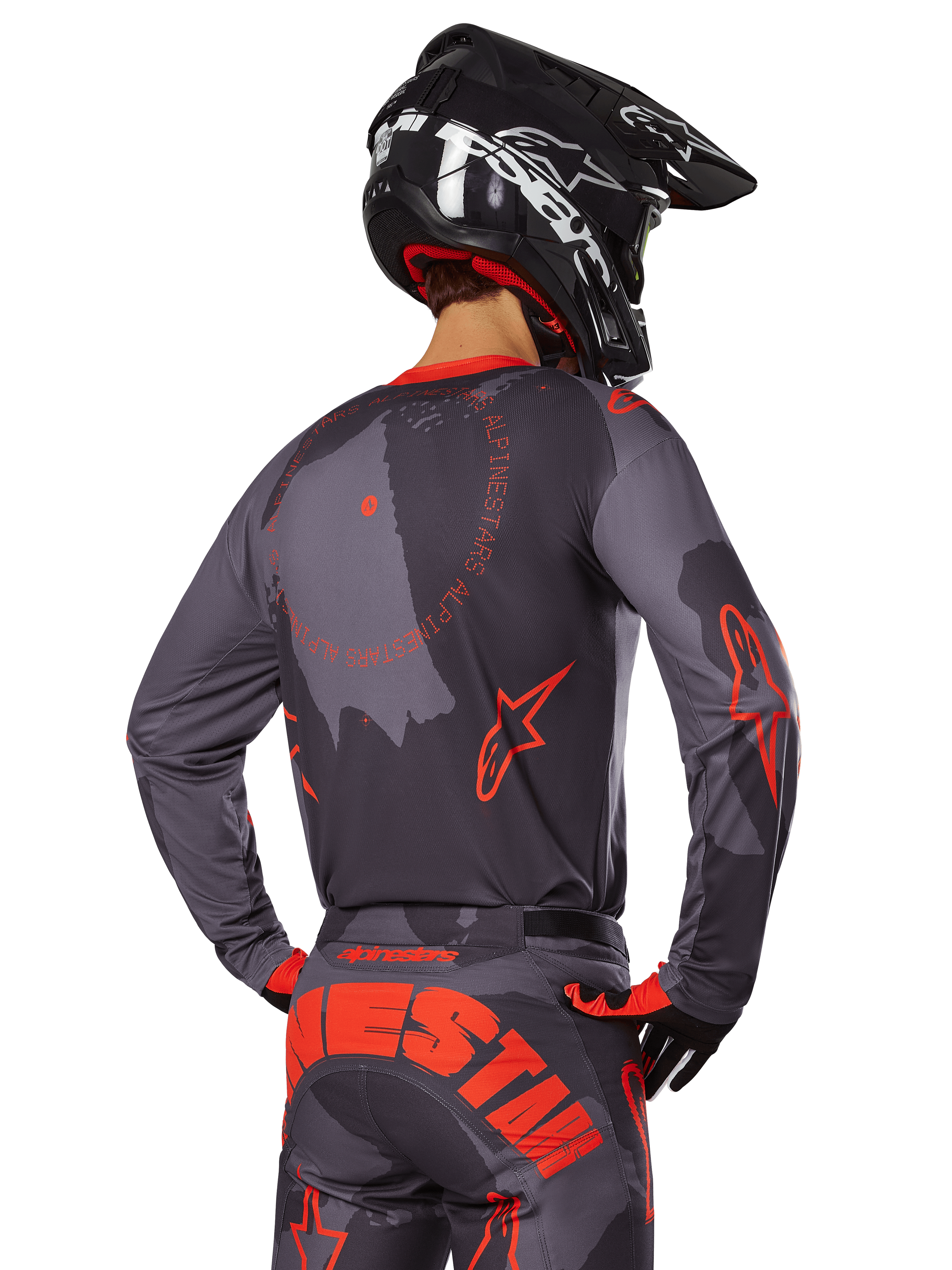 Zawodnik motocrossowy Alpinestars od tyłu w pełnym stroju wyścigowym, koszulka Racer Hollow w szarym kamuflażu z jaskrawopomarańczowymi akcentami i okrągłym logo na plecach, pasujące spodnie z dużym pomarańczowym napisem Alpinestars, czarny kask i pomarańczowo-czarne rękawice