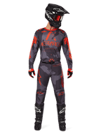 Zestaw Alpinestars 2025 Racer Hollow, zawodnik motocrossowy w koszulce i spodniach w szarym kamuflażu z jaskrawopomarańczowymi akcentami, czarny kask z pomarańczowymi goglami, czerwono-czarne rękawice, czarne buty z białym logo, pełny strój wyścigowy off-road