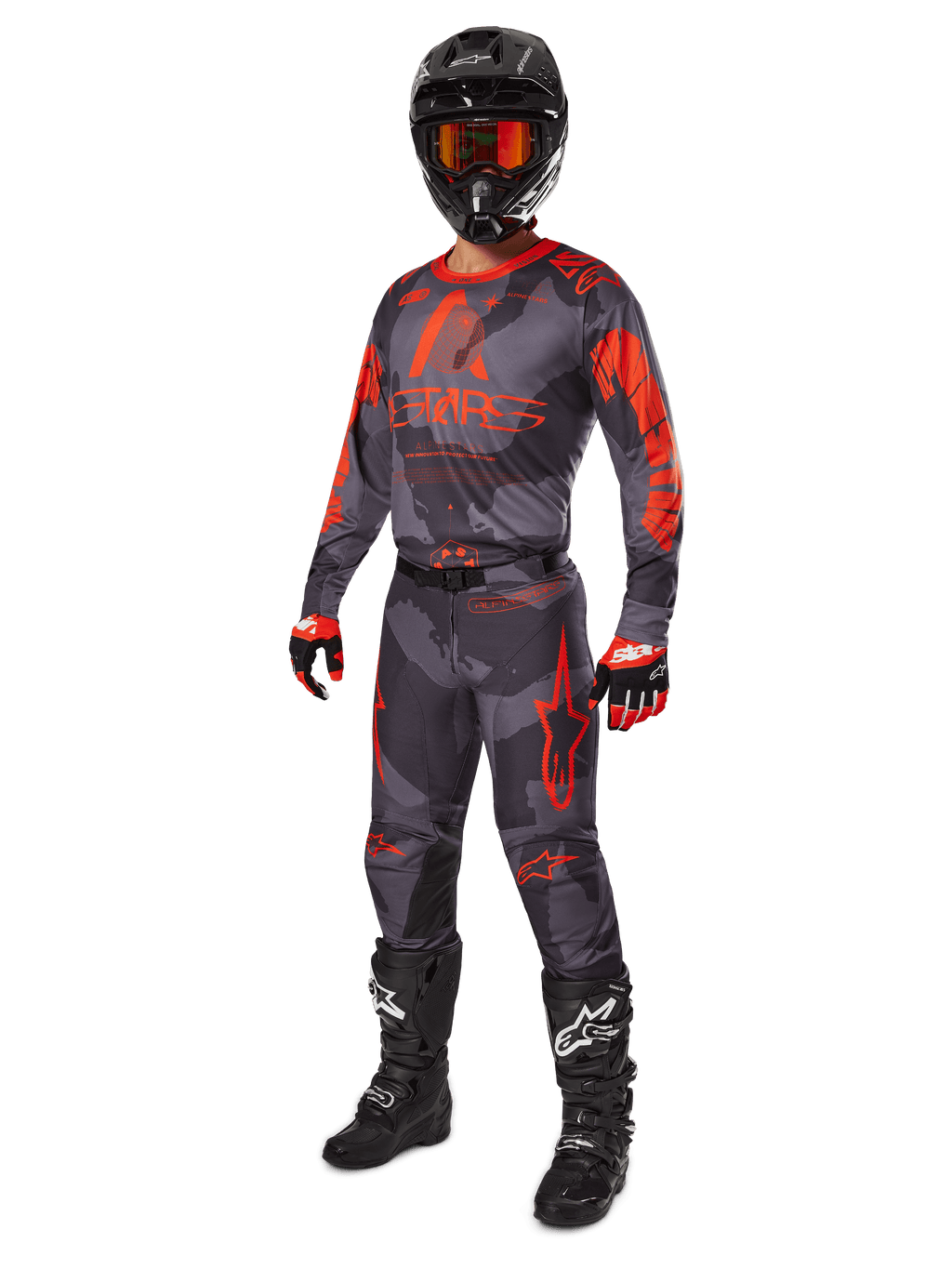 Zawodnik motocrossowy w pełnym stroju wyścigowym, koszulka i spodnie Alpinestars 2025 Racer Hollow w kolorze Gray Camo Orange Fluo, czarny kask z pomarańczowymi goglami, czarno-czerwone rękawice i czarne buty z białym logo, profesjonalny zestaw odzieży do jazdy terenowej