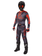 Zawodnik motocrossowy w pełnym stroju wyścigowym, koszulka i spodnie Alpinestars 2025 Racer Hollow w kolorze Gray Camo Orange Fluo, czarny kask z pomarańczowymi goglami, czarno-czerwone rękawice i czarne buty z białym logo, profesjonalny zestaw odzieży do jazdy terenowej