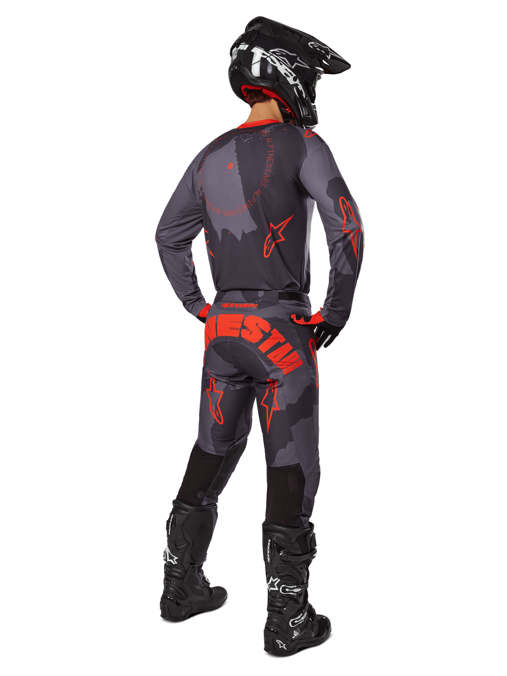 Zawodnik motocrossowy widziany z tyłu, ubrany w koszulkę Alpinestars 2025 Racer Hollow w kolorze Gray Camo Orange Fluo, pasujące spodnie camo z pomarańczowym napisem Alpinestars, czarny kask i czarne buty, dopasowany zestaw odzieży do jazdy terenowej